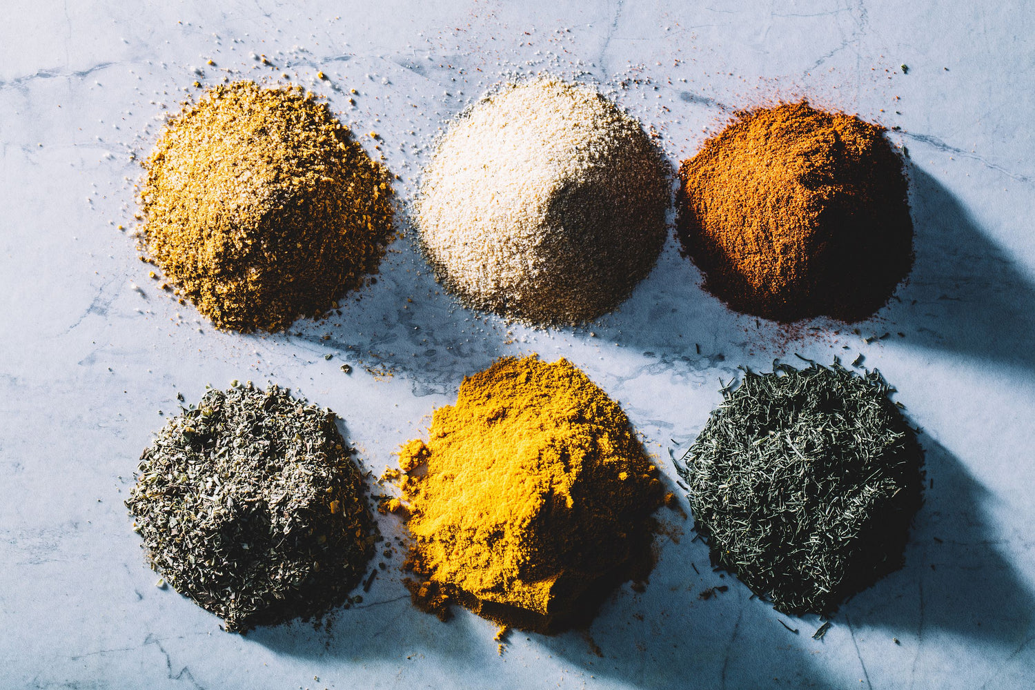 Six colorful piles of various ground spices and dried herbs on a light surface